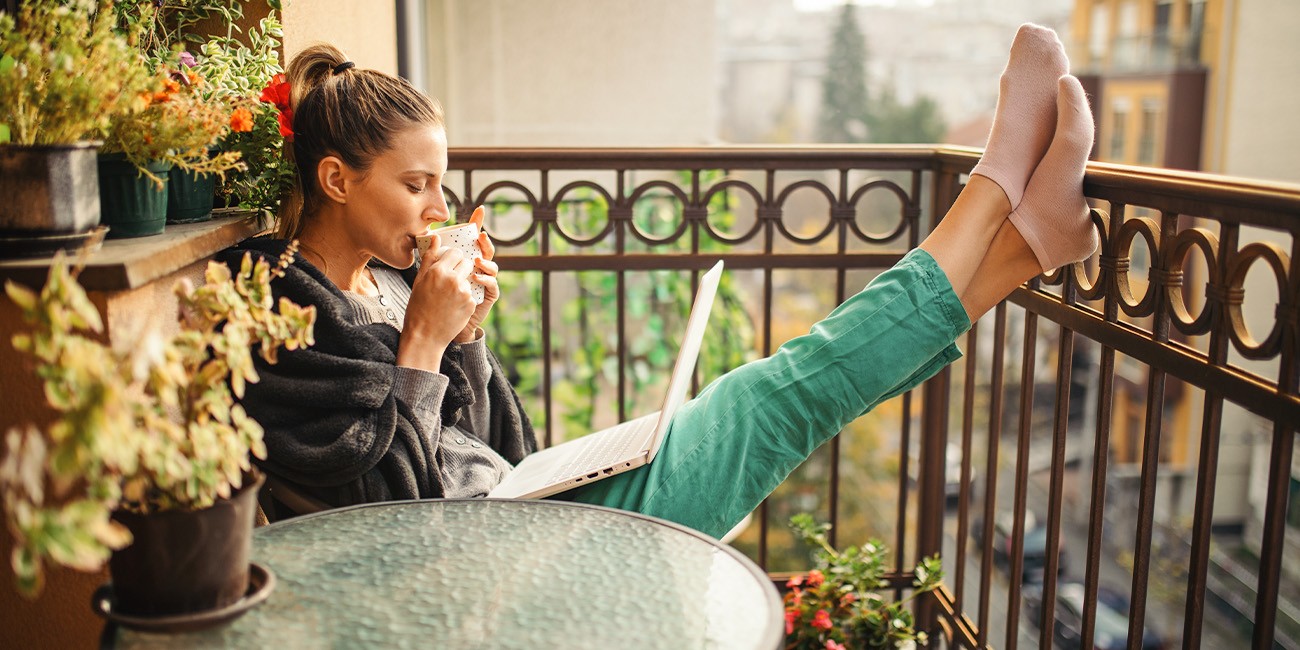 Frau sitzt entspannt auf Balkon mit Beinen hochgelegt und Laptop auf dem Schoß